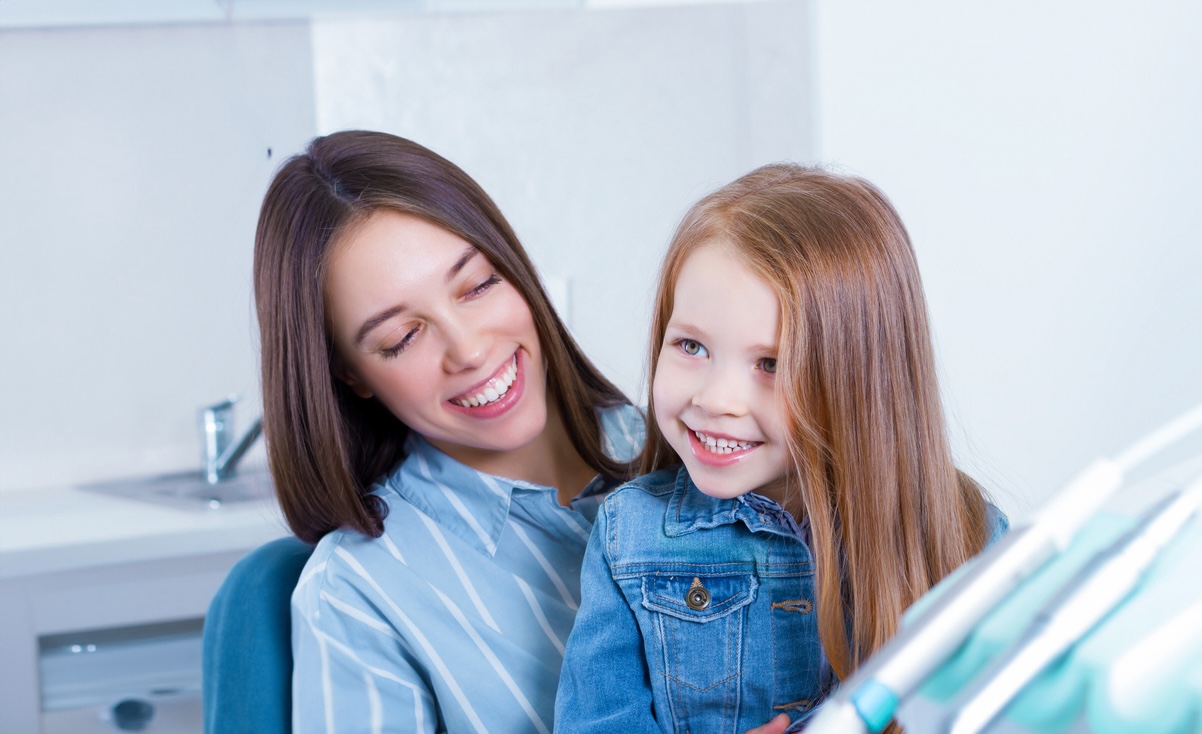 Girl in a jean jacket smiling up at her orthodontist from her mother’s lap