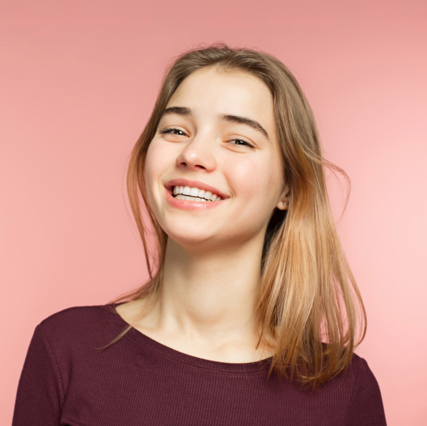 Woman smiling with perfect smile and white teeth on the pink studio background and looking at camera. Pretty young girl. She smiles widely. Her hair is light brown.